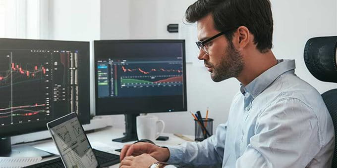 Un uomo con occhiali lavora su un laptop davanti a due monitor con grafici. Un uomo con occhiali lavora su un laptop davanti a due monitor con grafici.