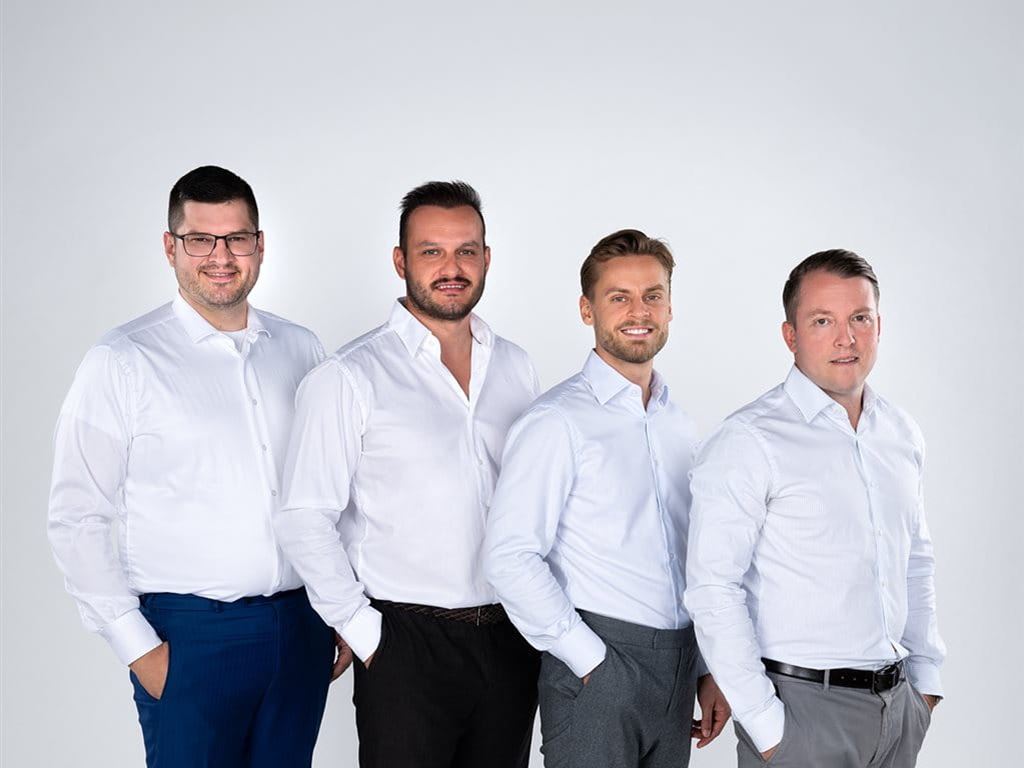 Vier Männer stehen nebeneinander, alle in weißen Hemden und Anzughosen, vor einem hellen Hintergrund.