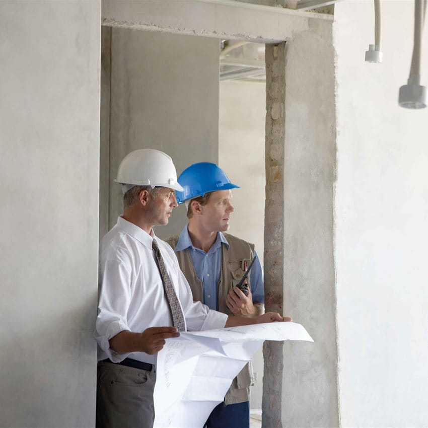 Due operai edili con caschi esaminano i piani di costruzione in uno spazio non finito. Due operai edili con caschi esaminano i piani di costruzione in uno spazio non finito.
