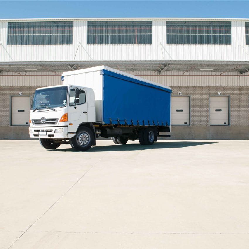 A white truck with a blue tarp is parked in front of a warehouse building with several doors. A white truck with a blue tarp is parked in front of a warehouse building with several doors.