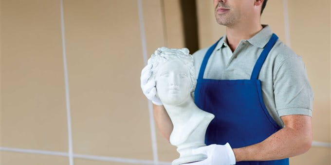 Eine Person in Arbeitskleidung hält eine weiße Statue in den Händen, im Hintergrund sind Kisten sichtbar. Eine Person in Arbeitskleidung hält eine weiße Statue in den Händen, im Hintergrund sind Kisten sichtbar.