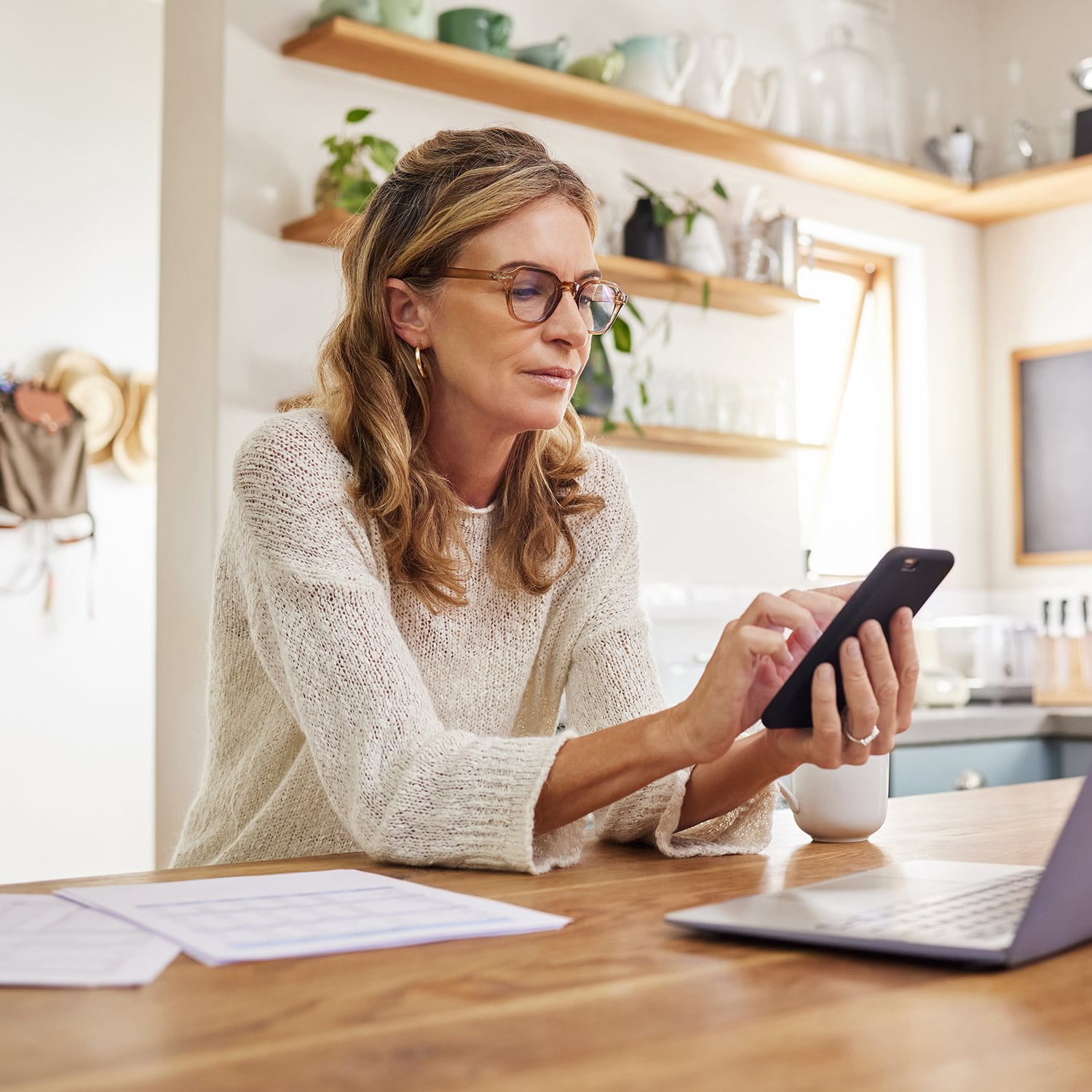 Frau sitzt am Küchentisch und nutzt Smartphone und Laptop, mit Unterlagen und einer Tasse vor sich.