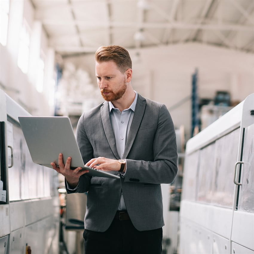 Ein Mann im Business-Outfit arbeitet mit einem Laptop in einer modernen Industriehalle, umgeben von grossen Maschinen. Ein Mann im Business-Outfit arbeitet mit einem Laptop in einer modernen Industriehalle, umgeben von grossen Maschinen.