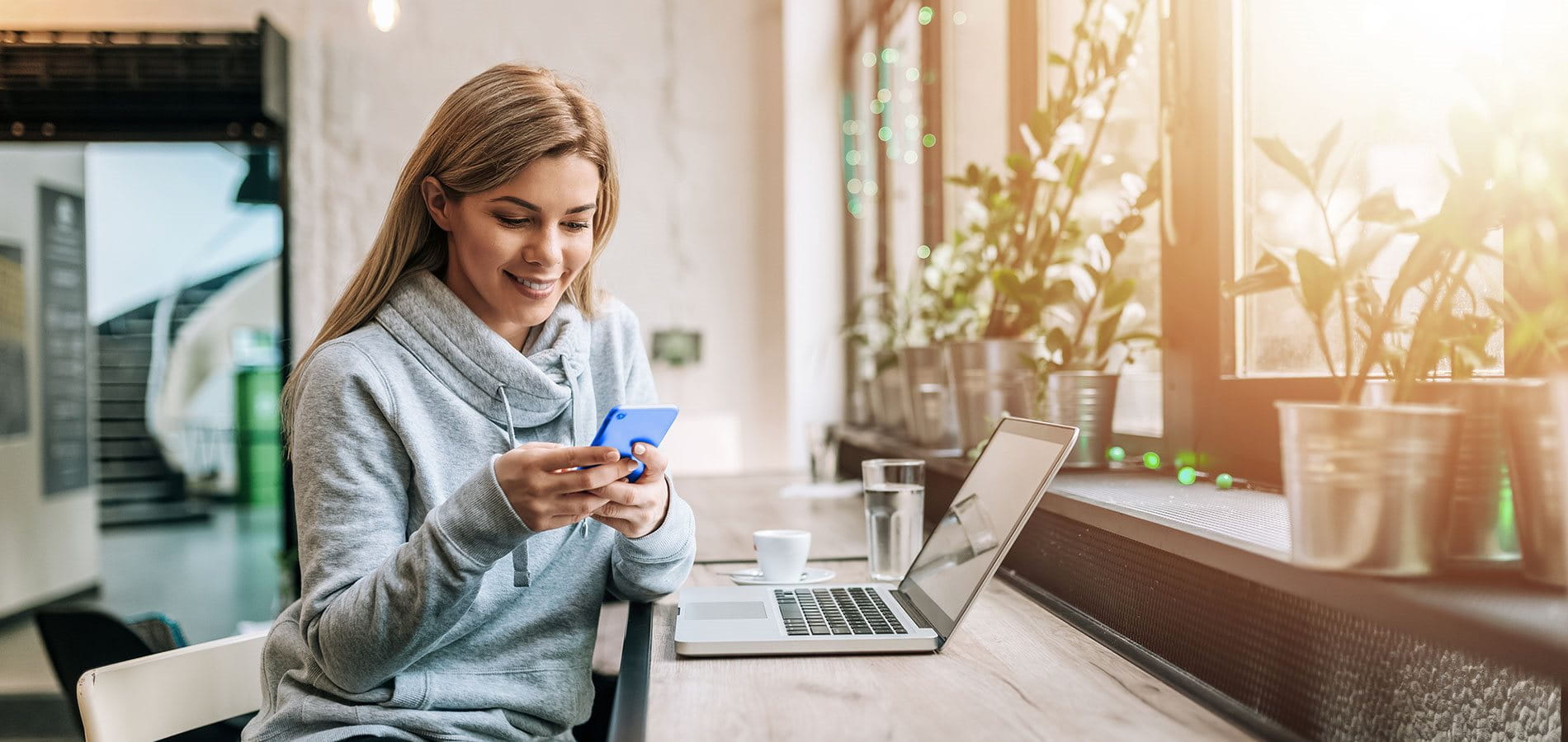 Eine junge Frau sitzt lächelnd an einem Fensterarbeitsplatz vor Ihrem Laptop und schaut auf ihr Mobile.