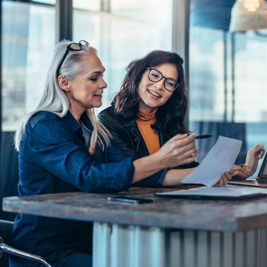 Zwei Frauen sitzen gemeinsam an einem Tisch in einem modernen Büro und besprechen Dokumente. Eine der Frauen hält einen Stift und zeigt auf ein Blatt Papier, während die andere aufmerksam zuhört. Zwei Frauen sitzen gemeinsam an einem Tisch in einem modernen Büro und besprechen Dokumente. Eine der Frauen hält einen Stift und zeigt auf ein Blatt Papier, während die andere aufmerksam zuhört.