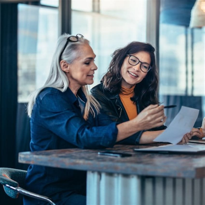 Zwei Frauen sitzen gemeinsam an einem Tisch in einem modernen Büro und besprechen Dokumente. Eine der Frauen hält einen Stift und zeigt auf ein Blatt Papier, während die andere aufmerksam zuhört. Zwei Frauen sitzen gemeinsam an einem Tisch in einem modernen Büro und besprechen Dokumente. Eine der Frauen hält einen Stift und zeigt auf ein Blatt Papier, während die andere aufmerksam zuhört.