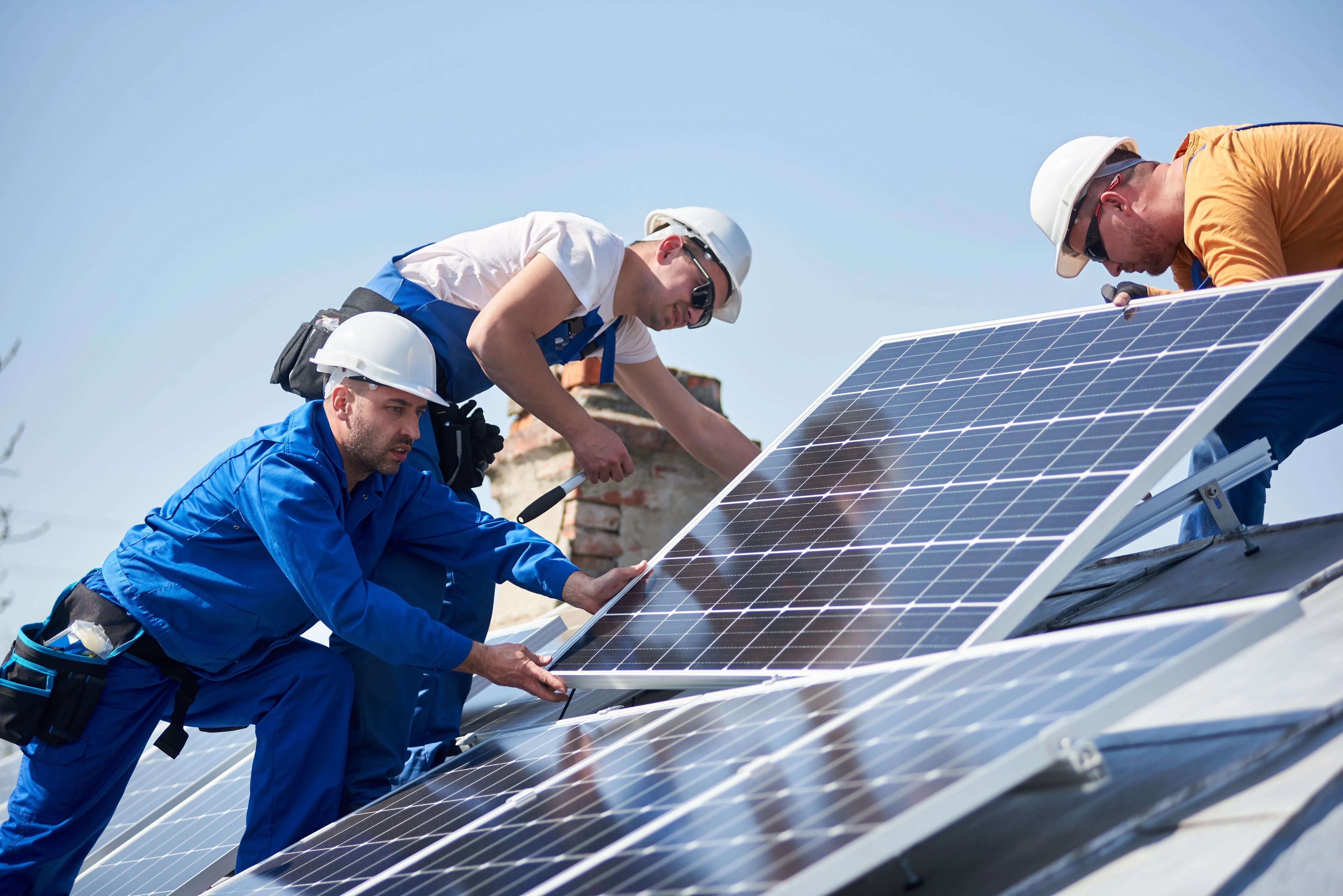 Drei Arbeiter installieren Solarpanels auf einem Dach unter blauem Himmel.