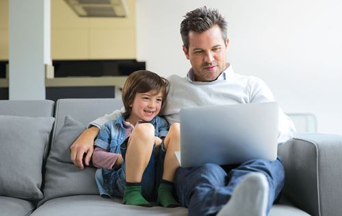Ein Mann und ein Kind sitzen auf einem Sofa und schauen auf einen Laptop mit Freude.