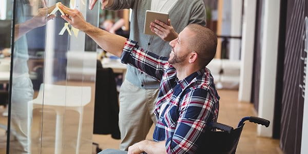 Au bureau, un homme en fauteuil roulant esquisse une idée avec son collègue de travail sur une paroi en verre.