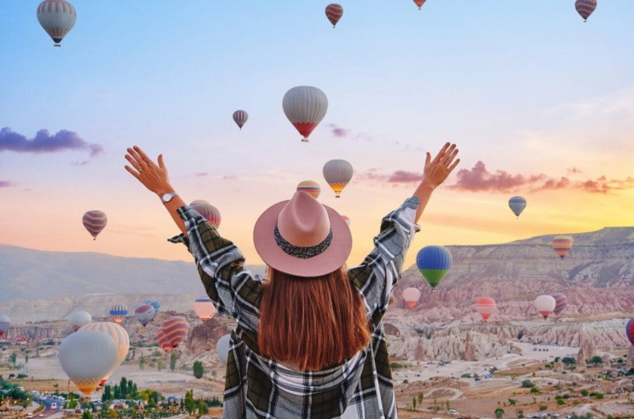 Frau mit erhobenen Armen betrachtet viele Heissluftballons am Himmel bei Sonnenaufgang.