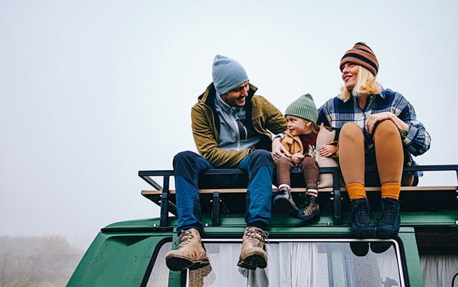 An adult and a child sit together at a breakfast table in a bright kitchen. In front of them are bowls of cereal, glasses of orange juice, mugs, a plate of bread slices, and a bowl of fresh fruit. Both have their arms raised and appear to be interacting in a joyful manner.