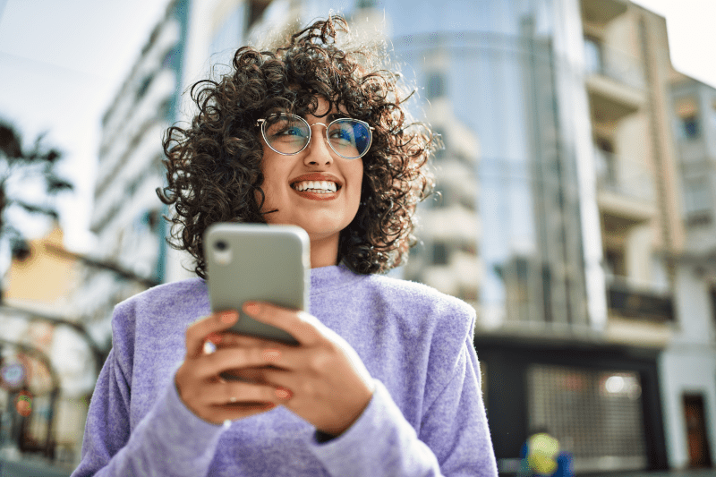 Eine Person mit lockigen Haaren steht draußen in einer Stadt und hält ein Smartphone in beiden Händen. Sie trägt einen lila Pullover. Im Hintergrund sind moderne Gebäude und Glasfassaden zu sehen.
