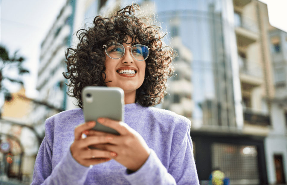 Una persona con capelli ricci si trova all’aperto in città e tiene uno smartphone con entrambe le mani. Indossa un maglione viola. Sullo sfondo si vedono edifici moderni con facciate di vetro. Una persona con capelli ricci si trova all’aperto in città e tiene uno smartphone con entrambe le mani. Indossa un maglione viola. Sullo sfondo si vedono edifici moderni con facciate di vetro.