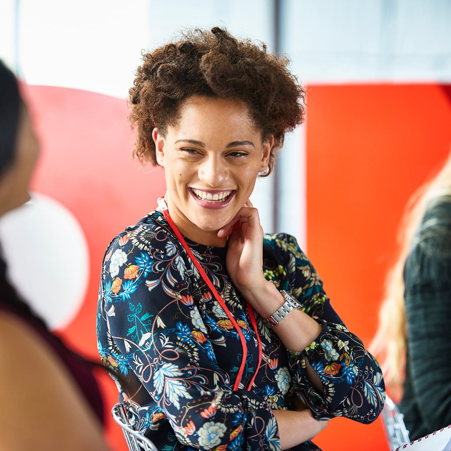 Person with curly hair and colorful blouse talking to two others in front of a red background.