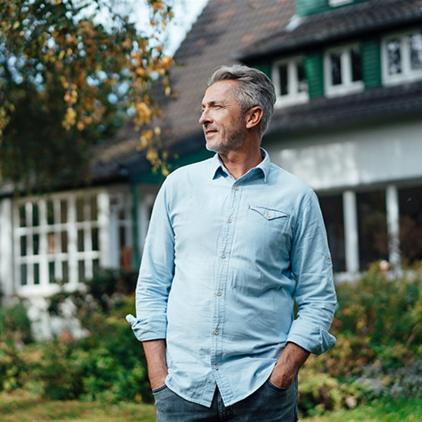 A man stands with his hands in his pockets in front of a residential house with green shutters. He is wearing a light blue shirt and is looking to the left. The background features a garden and trees with autumn-colored leaves. A man stands with his hands in his pockets in front of a residential house with green shutters. He is wearing a light blue shirt and is looking to the left. The background features a garden and trees with autumn-colored leaves.