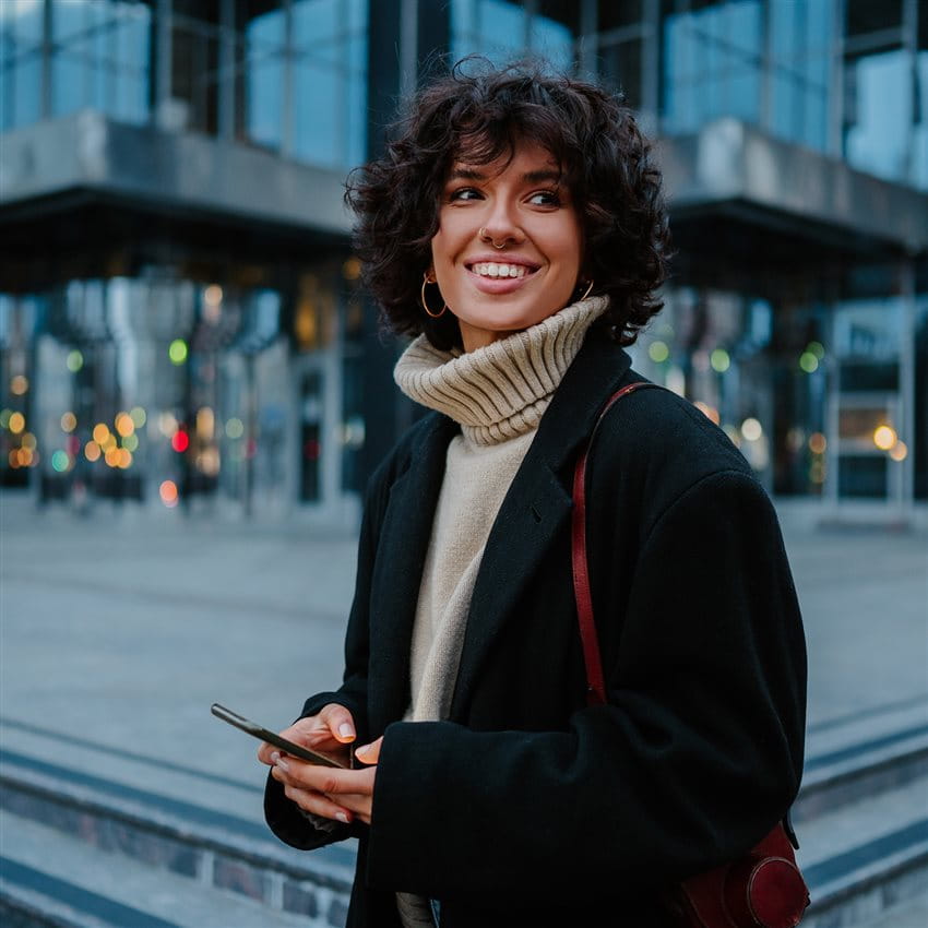 Persona con capelli ricci, cappotto scuro e smartphone davanti a un edificio. Persona con capelli ricci, cappotto scuro e smartphone davanti a un edificio.