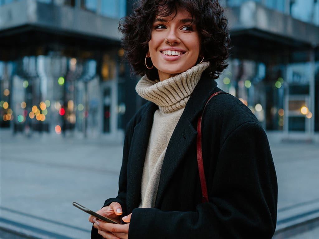 Persona con capelli ricci, cappotto scuro e smartphone davanti a un edificio.