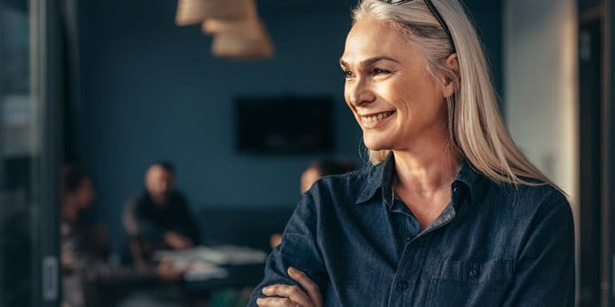 A woman with long light hair stands with her arms crossed and glasses on her head in a modern office. In the background, people are sitting at a table. A woman with long light hair stands with her arms crossed and glasses on her head in a modern office. In the background, people are sitting at a table.