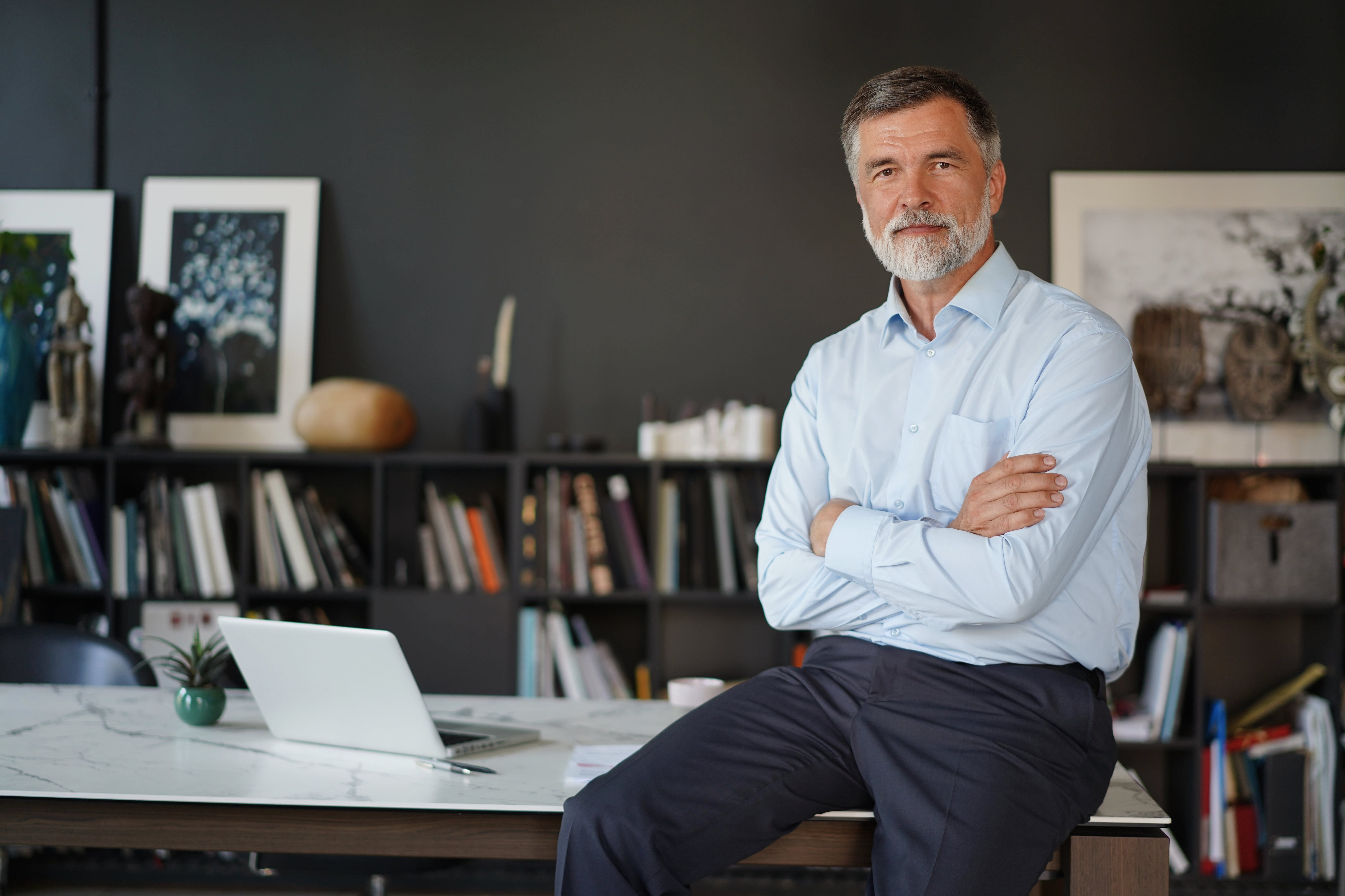 A man in light clothing sits with his arms crossed on a desk in a modern office with shelves, books, and a laptop.