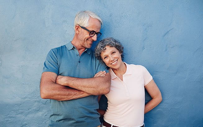 Ein älteres Paar steht vor einer blauen Wand, lehnt sich entspannt aneinander und trägt sommerliche Kleidung mit verschränkten Armen.