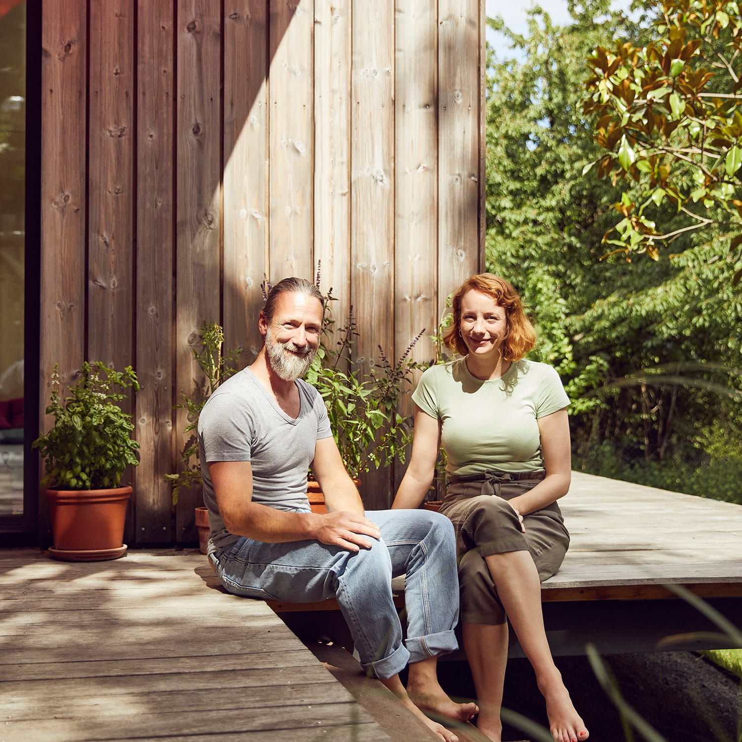 A couple sits barefoot on a wooden terrace in front of a modern wooden house, surrounded by plants.