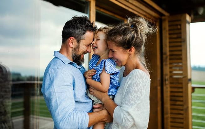 A young family stands closely together on a terrace in front of a house, enjoying time outdoors as a family.
