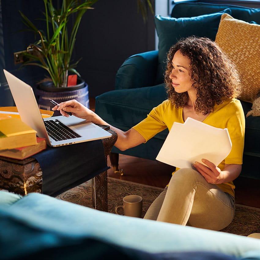 A person sits with a laptop, holding papers and a pen in hand. A person sits with a laptop, holding papers and a pen in hand.