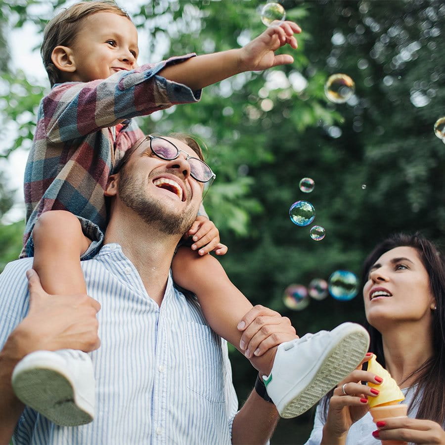 A child sitting on his father's shoulders grabs soap bubbles created by his mother.
