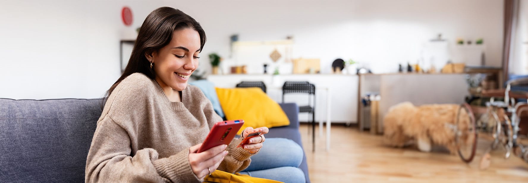 Frau sitzt lächelnd auf einem Sofa im Wohnzimmer, hält eine Kreditkarte und schaut auf ihr Smartphone.