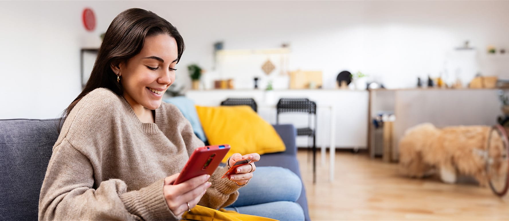Frau sitzt lächelnd auf einem Sofa im Wohnzimmer, hält eine Kreditkarte und schaut auf ihr Smartphone. Frau sitzt lächelnd auf einem Sofa im Wohnzimmer, hält eine Kreditkarte und schaut auf ihr Smartphone.