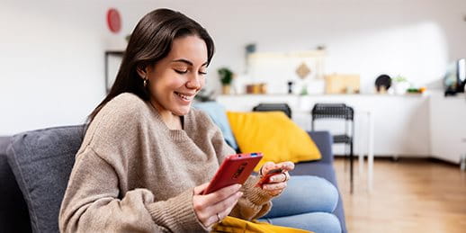 Frau sitzt lächelnd auf einem Sofa im Wohnzimmer, hält eine Kreditkarte und schaut auf ihr Smartphone.