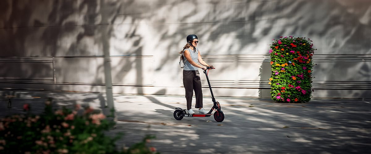 A person rides an electric scooter in a quiet area with flowers. They are wearing a helmet.