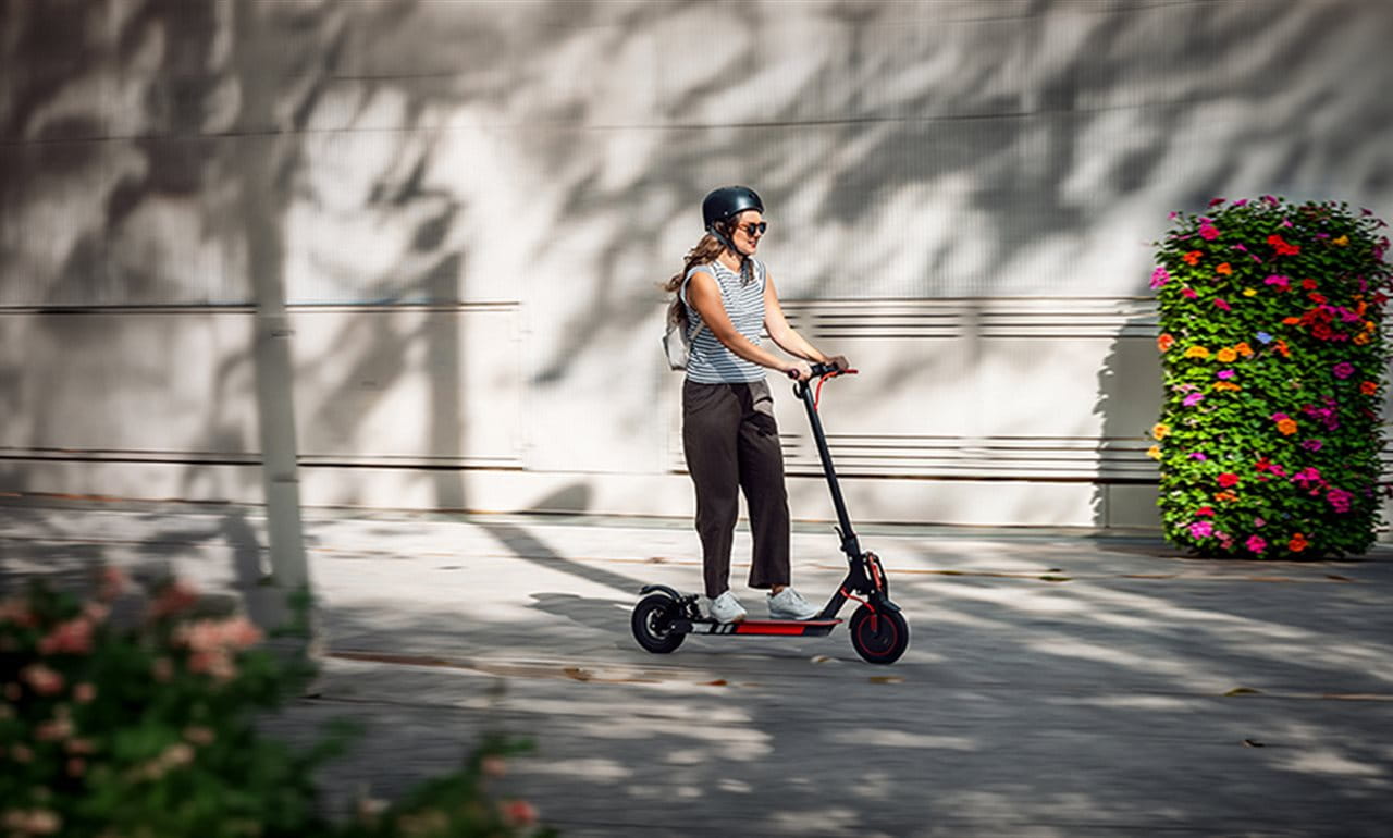 A person rides an electric scooter in a quiet area with flowers. They are wearing a helmet. A person rides an electric scooter in a quiet area with flowers. They are wearing a helmet.