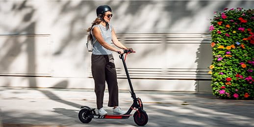 Une personne roule sur un trottinette. À l'arrière-plan, il y a un parterre de fleurs.