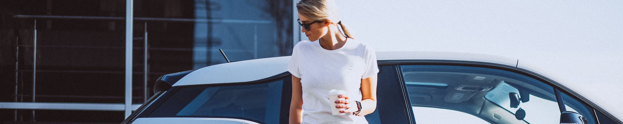  Woman stands in front of her car at the electric charging station and drinks coffee