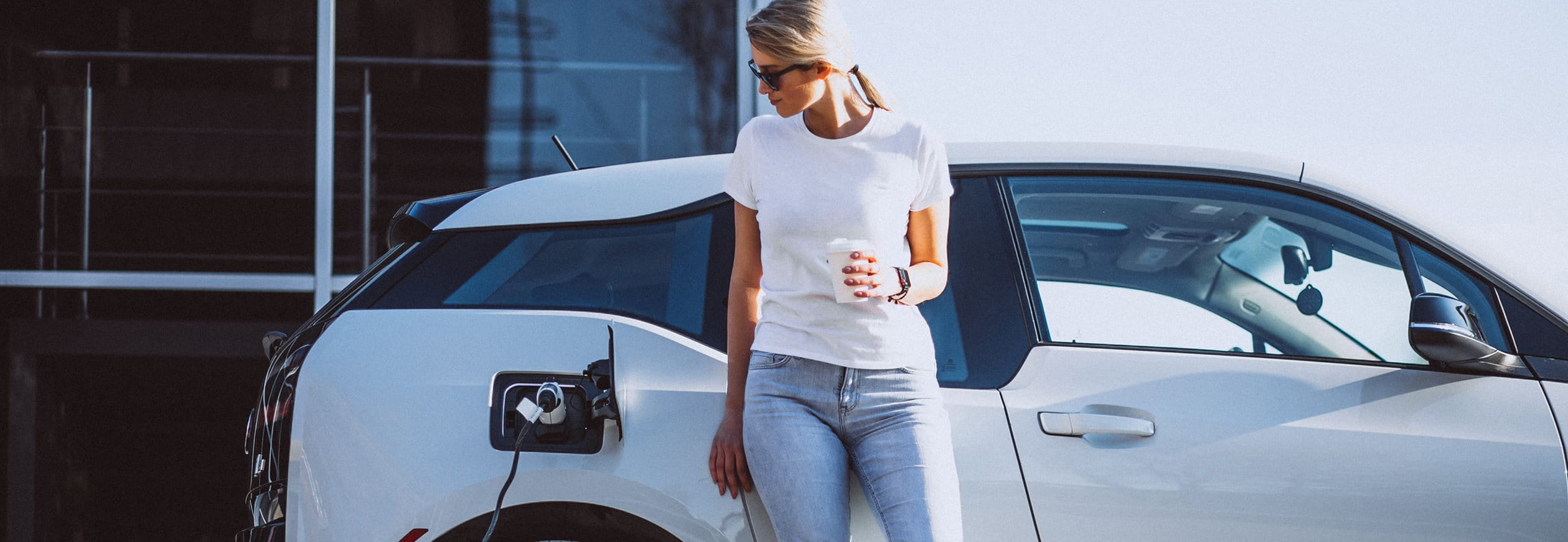Une femme se tient devant sa voiture à la station de charge électrique et boit du café