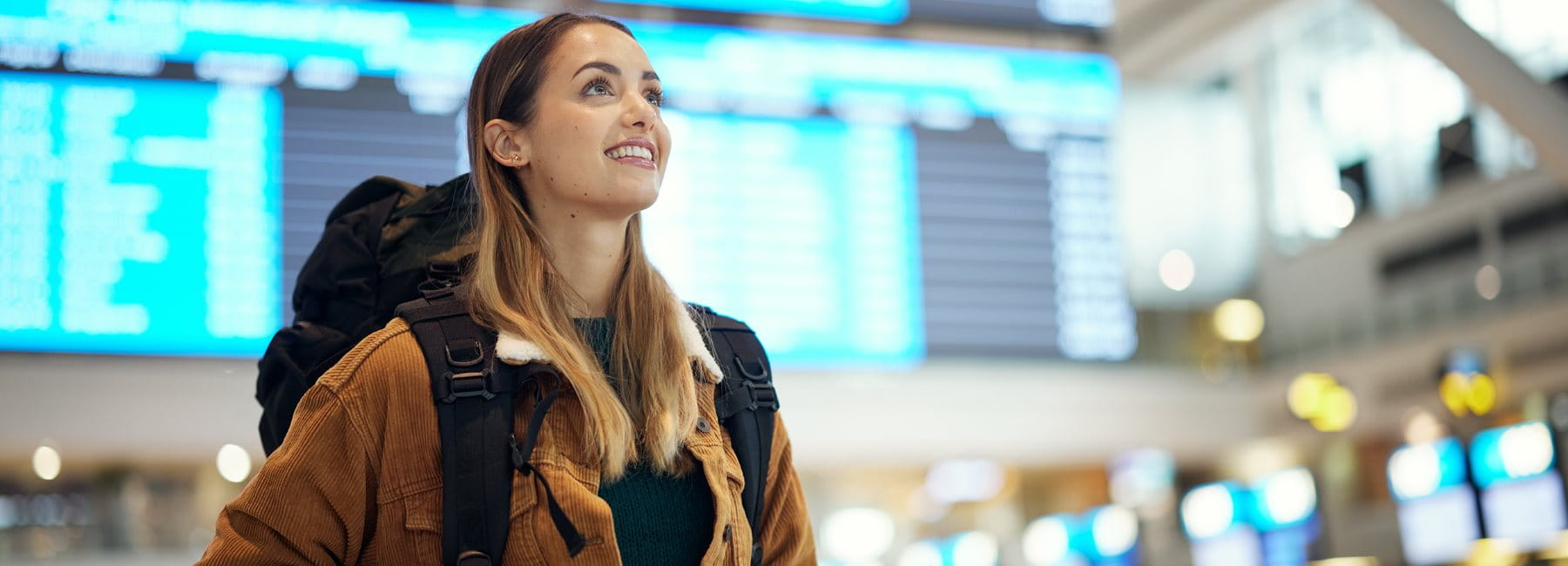 Eine junge Frau mit Rucksack steht am Flughafen und hält Reisedokumente in der Hand. Im Hintergrund sind Abflugtafeln und das geschäftige Flughafenumfeld zu sehen.