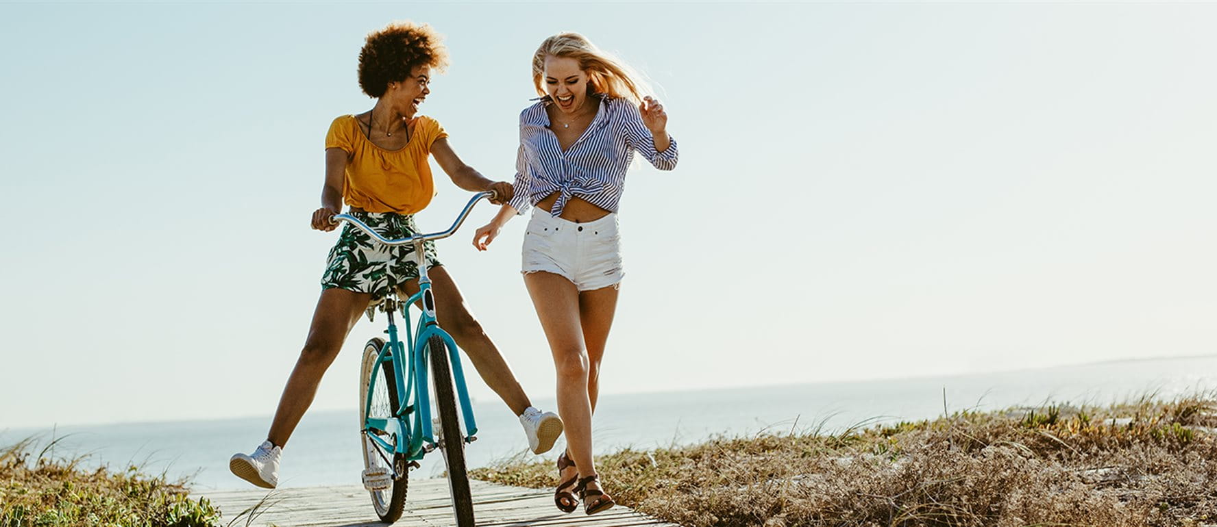 Zwei junge Frauen am Strand – eine fährt lächelnd Fahrrad, die andere läuft nebenher. Zwei junge Frauen am Strand – eine fährt lächelnd Fahrrad, die andere läuft nebenher.
