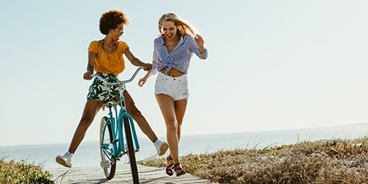 Two young women at the beach – one is riding a bike and smiling, the other is walking alongside. Two young women at the beach – one is riding a bike and smiling, the other is walking alongside.