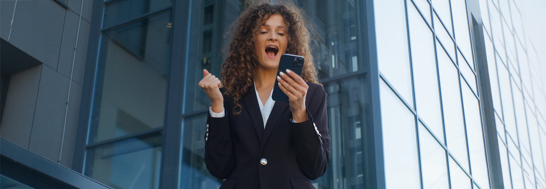 Una donna in giacca tiene uno smartphone e sorride entusiasta davanti a un moderno edificio.