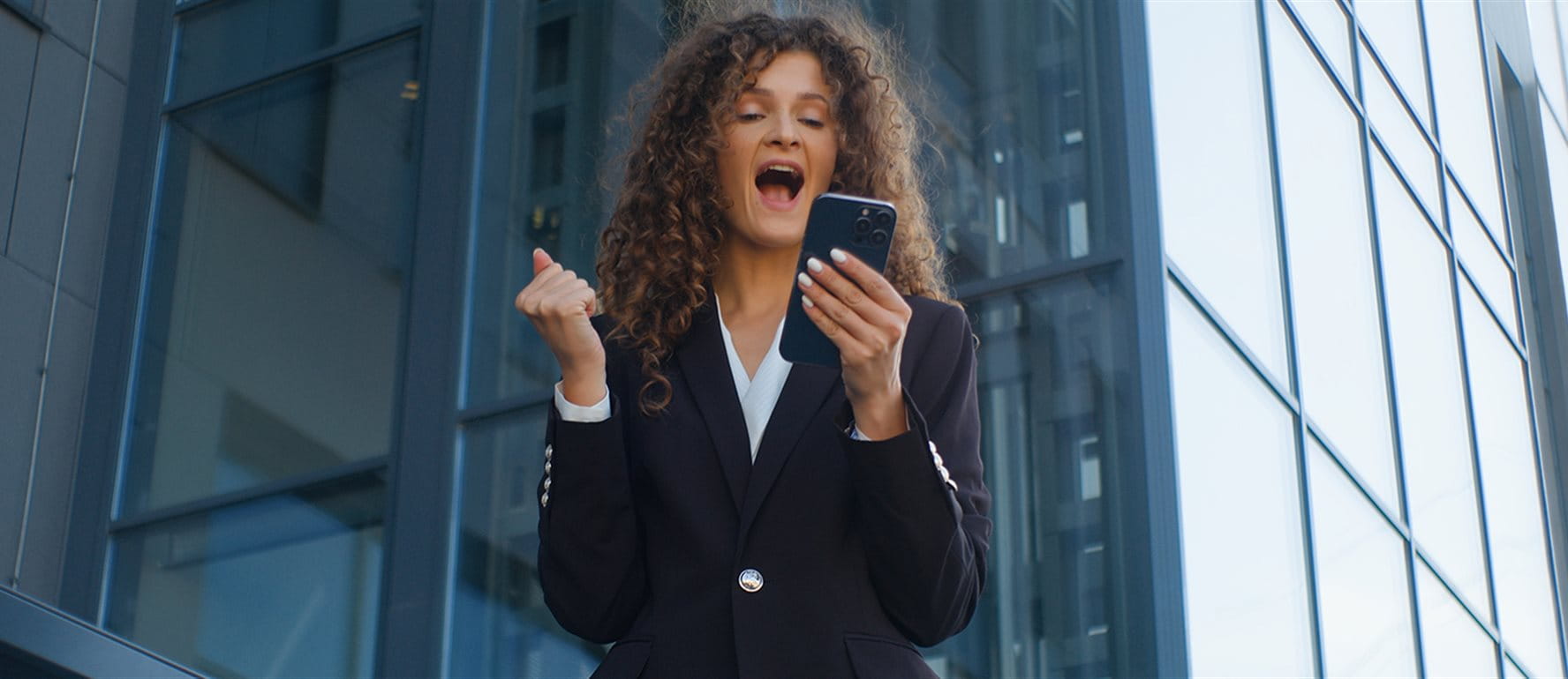 Una donna in giacca tiene uno smartphone e sorride entusiasta davanti a un moderno edificio. Una donna in giacca tiene uno smartphone e sorride entusiasta davanti a un moderno edificio.