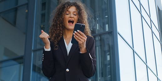 Una persona in abito tiene un cellulare e mostra gioia. Sullo sfondo si vedono edifici moderni.