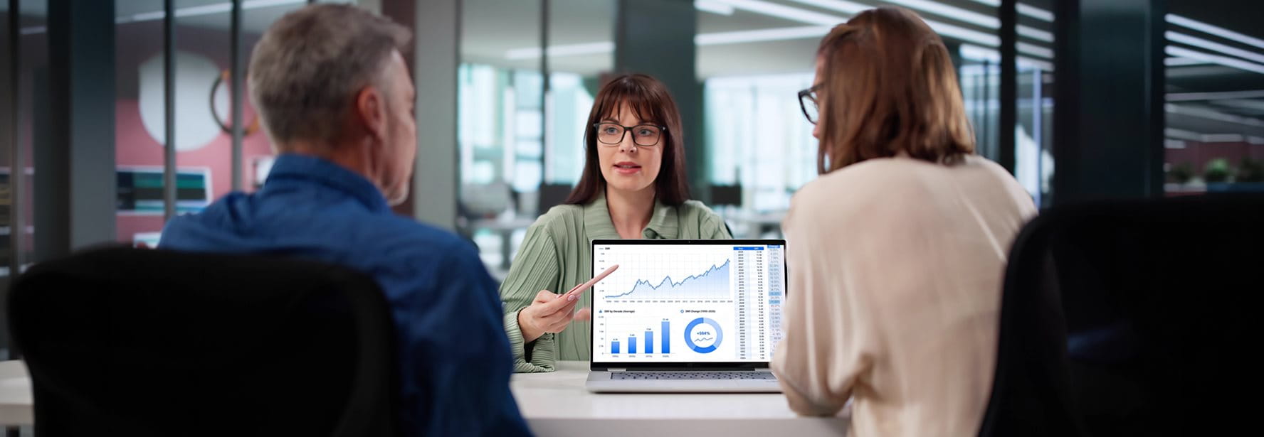 Three people in a meeting room, a presentation on a laptop shows charts and data.
