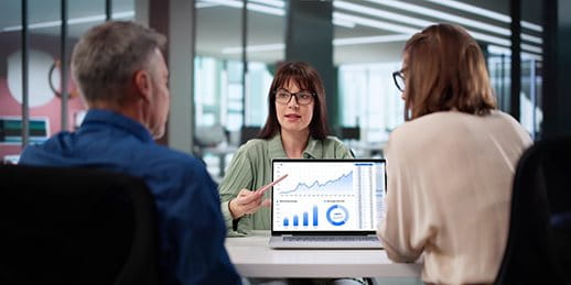 Three people are discussing in a modern office. One person points at a graph on a laptop.