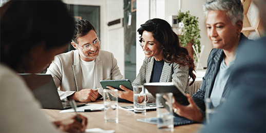 A group of people sit around a table during a meeting, engaging with digital devices and papers. A group of people sit around a table during a meeting, engaging with digital devices and papers.