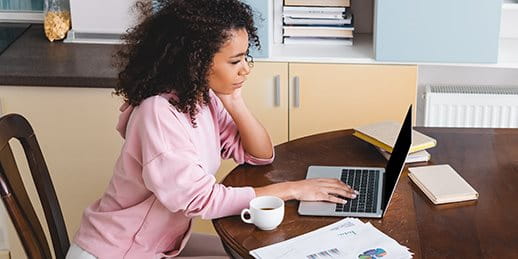 Une femme assise à la table de la cuisine travaille sur un ordinateur portable, des documents fiscaux sont posés à côté d’elle.