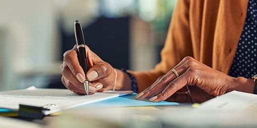 A close-up of a person's hand writing with a pen on a piece of paper, surrounded by documents and stationery.