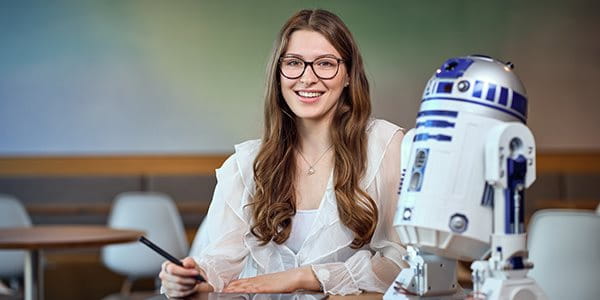 Una persona è seduta a un tavolo in una stanza moderna e lavora con un tablet e una penna. Accanto a lei c’è un modello del robot R2-D2 di Star Wars.