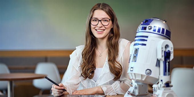 Una persona è seduta a un tavolo in una stanza moderna e lavora con un tablet e una penna. Accanto a lei c’è un modello del robot R2-D2 di Star Wars. Una persona è seduta a un tavolo in una stanza moderna e lavora con un tablet e una penna. Accanto a lei c’è un modello del robot R2-D2 di Star Wars.