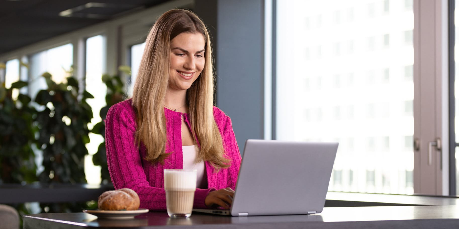 Eine Person mit langen Haaren arbeitet an einem Laptop mit einem Getränk und einem Brötchen.